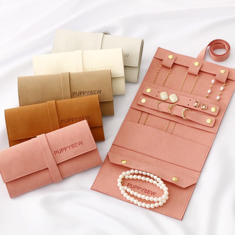 May include: A collection of jewelry travel cases in various colors, including pink, tan, and brown. The cases are made of a soft material and feature a strap closure. One case is open, displaying jewelry such as necklaces, earrings, and a pearl bracelet. The text "PUPPYSEW" is visible.